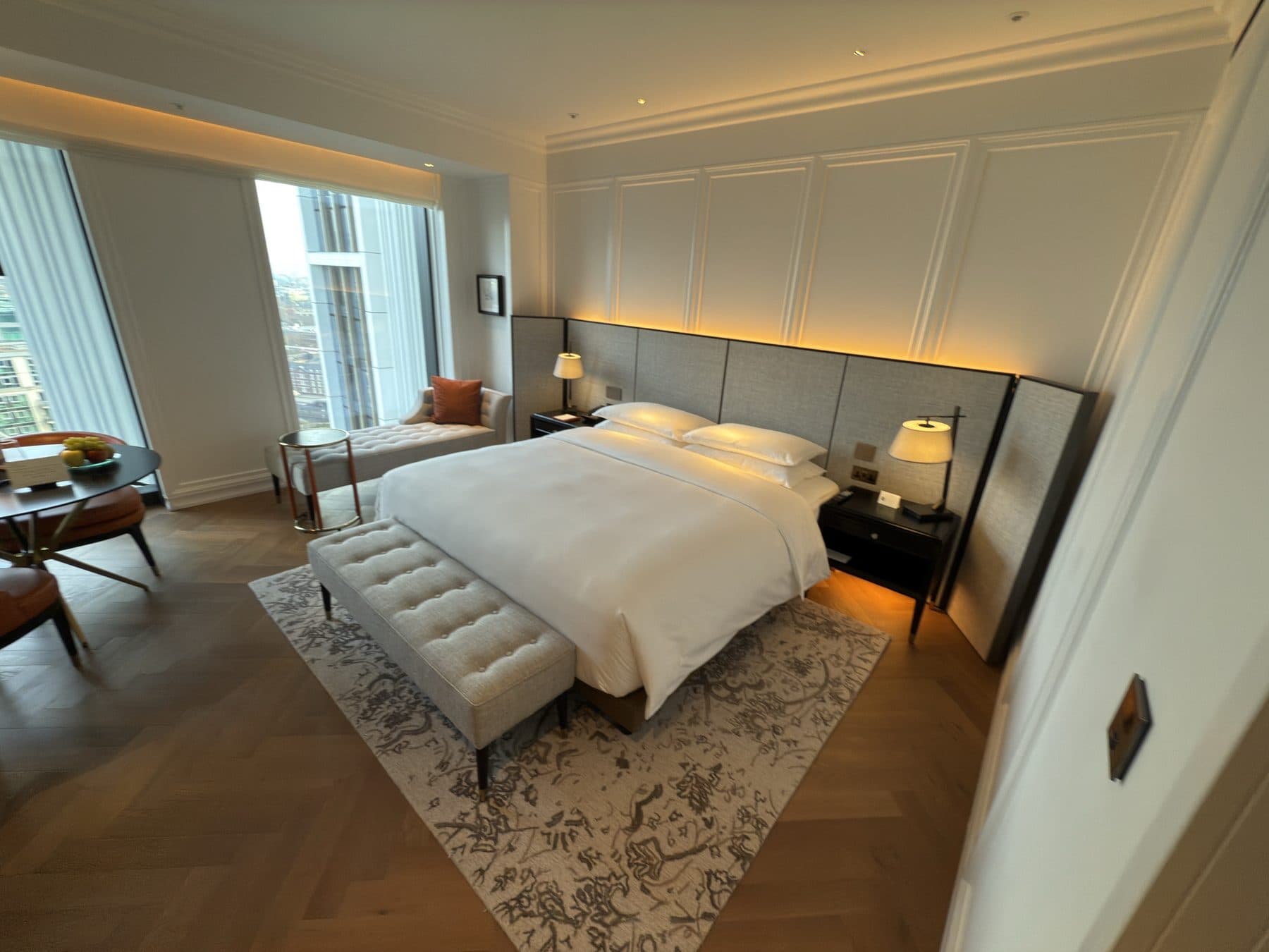 King room at a London hotel: white bed against a backlit fabric headboard, herringbone wood floor, and a chaise by tall windows overlooking the city